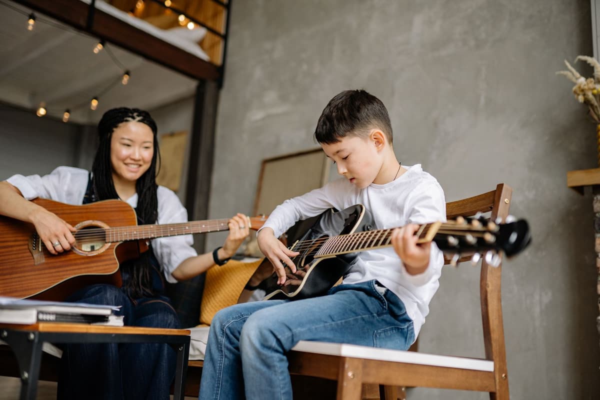 Ein Bub greift einen Akkord auf einer Gitarre, daneben spielt eine Lehrerin mit.